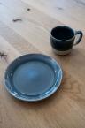 Blau glasierter Keramikteller und passender Becher mit Henkel auf heller Holztischplatte