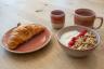 Frühstücksgedeck aus rosé-rotem Keramikgeschirr auf Holztisch: Teller mit Croissant, Becher, Tasse und Schale mit Joghurt, Müsli und Himbeeren