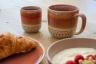 Keramikbecher und Tasse in Rot-Beige mit unglasiertem Dekorrand, dazu Teller mit Croissant und Schale mit Joghurt und Himbeeren auf Holztisch