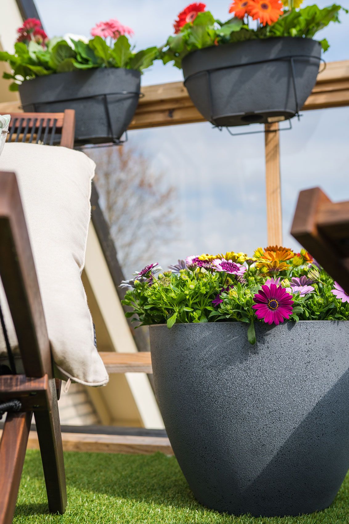 Terrasse mit einem großen bepflanzten Topf in dunkler Beton-Optik, darüber hängen zwei Balkonkästen an einer Pergola